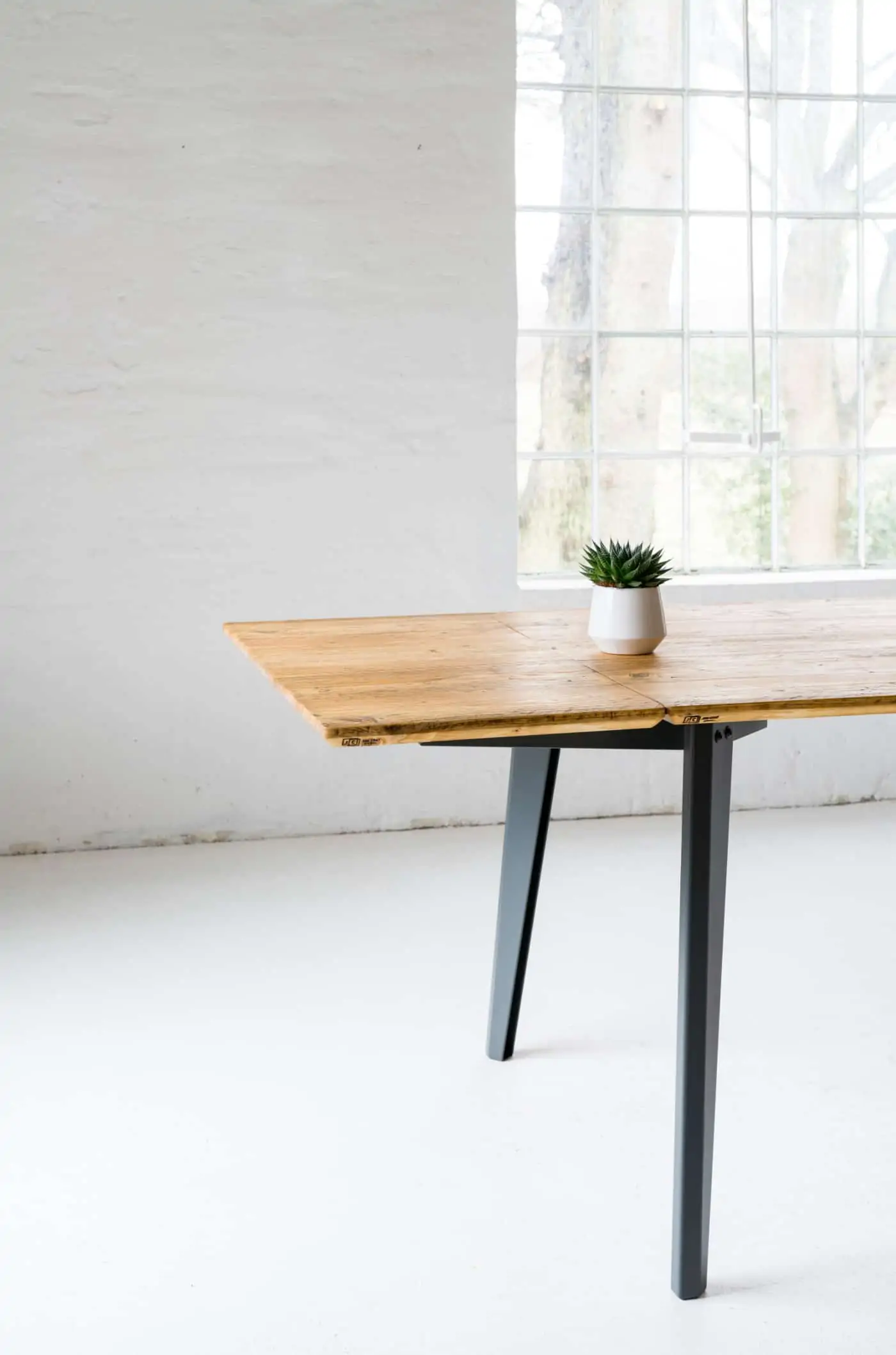 Esstisch mit Ansteckplatte aus recyceltem Bauholz und dunkelgrauem Metallgestell vor weißer Wand mit großzügigem Fenster