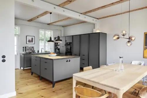 Blick in einen offenen Küchenbereich eines sanierten Altbaus. Eine freistehende Modulküche von Jan Cray aus Hamburg mit Inselmodul und Wandmodul mit jeweils dunklen Linoleumfronten sowie ein heller Holztisch und Lichtobjekten machen den Wohnraum zu einem wohnlichen Zuhause mit skandinavischem Ambiente.