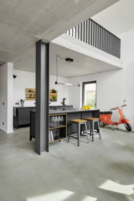 Der Wohn- und Kochbereich eines Lofts im Industriedesign. Stahlträger, Betonwände und ein gegossener Estrich zieren den Raum. Im Mittelpunkt des Raumes steht eine freistehende Modulküche aus Linoleum und Stahl. Ebenso ein Holztisch mit schwarzen Stühlen.